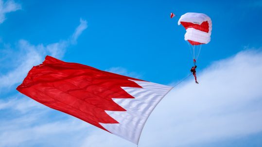 Skydiver, at the Bahrain International Airshow, falling through the air with parachute opened while holding a big Bahrain flag.