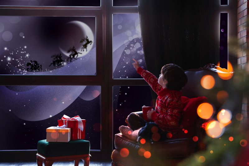 Little boy in red pajamas looking out of the window and seeing Santa Claus' reindeer flying on a snowy Christmas Eve night.