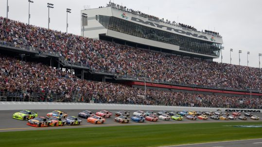 NASCAR cars racing in front of the grandstand filled with fans at the Daytona 500.