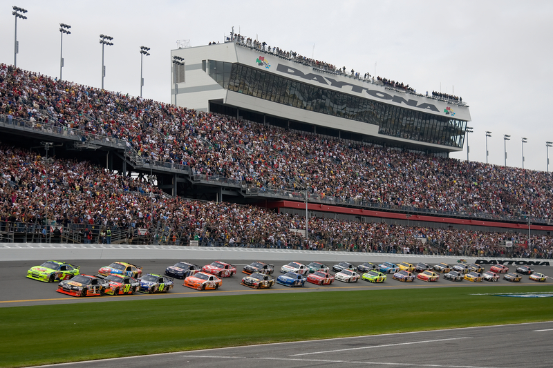 NASCAR cars racing in front of the grandstand filled with fans at the Daytona 500.