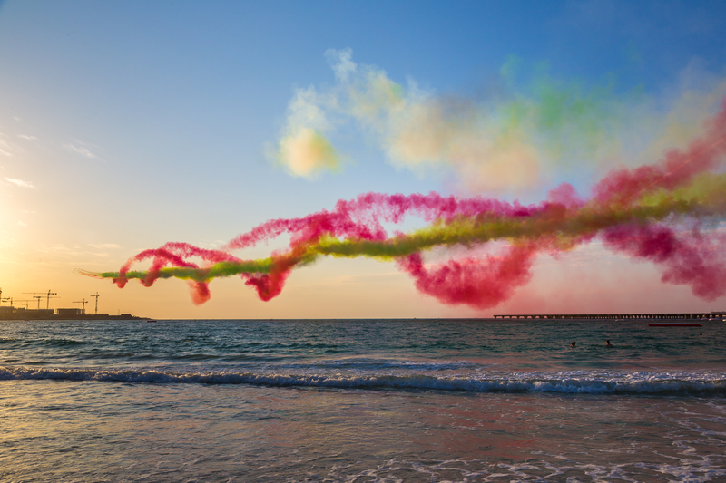 Airplanes performing stunts over the sea with red and green smoke trailing at the Dubai Airshow.