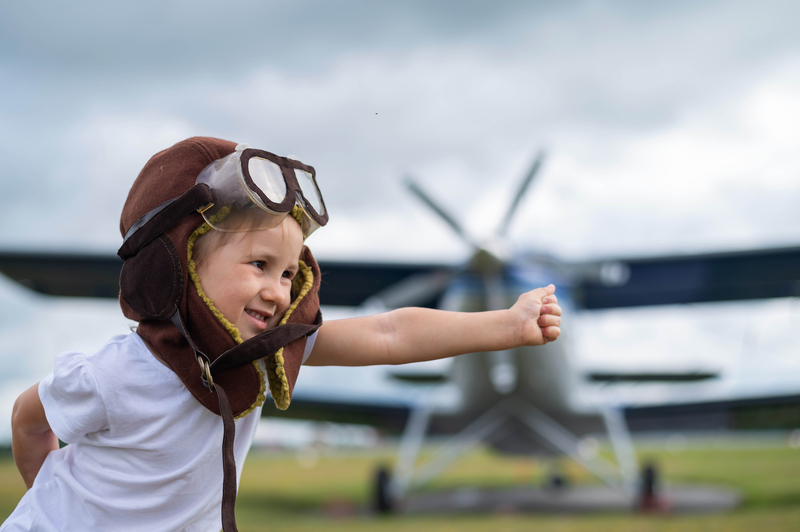 Little girl wearing an old pilot cap & goggles sticking her thumb up in front of a biplane on Girls in Aviation Day.
