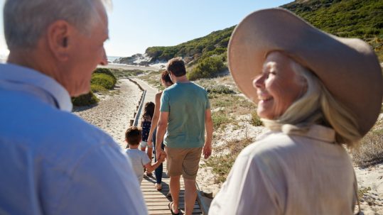 Grandmother and grandfather smiling at each other as they walk behind their family at the beach on Grandparents Day.