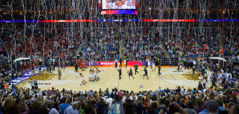 Basketball team and fans in the stadium celebrating while streamers come down from the rafters after winning the NCAA Women’s Final Four.