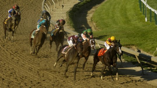 Horses & jockeys racing around the curve on the dirt track of the Preakness Stakes.