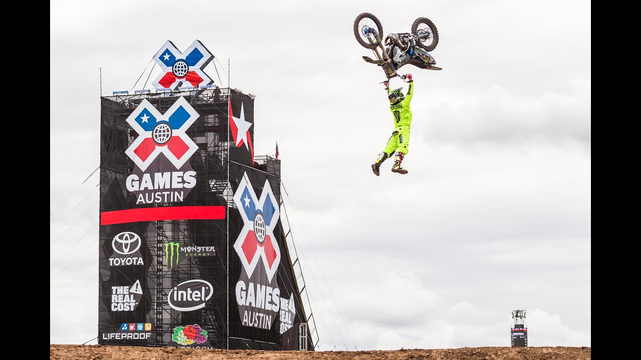 Motorcycle rider hanging from the handles underneath his upside-down motorcycle during a flip trick in front of a Summer X Games sign.