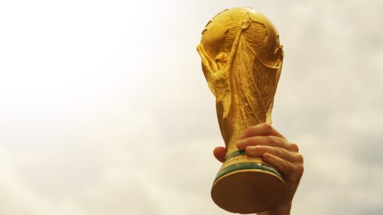 Hand raising the golden FIFA World Cup trophy into a sun-soaked sky.