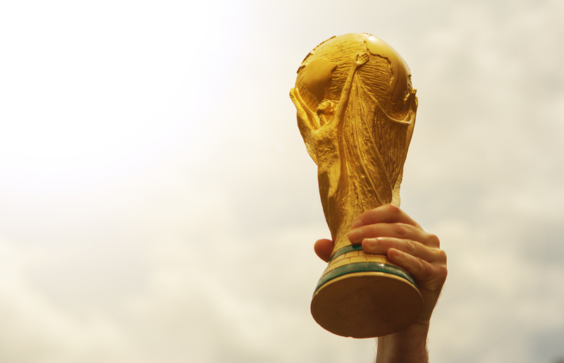 Hand raising the golden FIFA World Cup trophy into a sun-soaked sky.