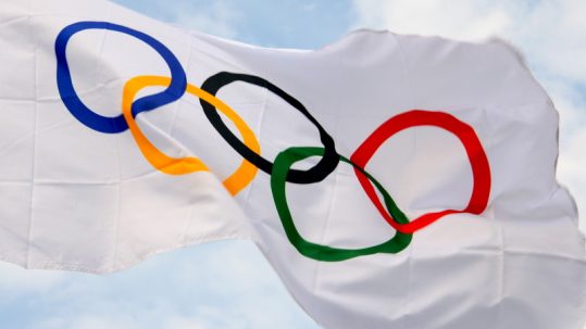 Flag with the five Olympic Rings waving in front of a blue and partly cloudy sky during the Summer Olympics.
