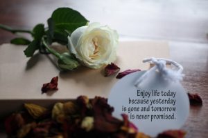 White rose and red rose petals laying on messy table with inspirational quote: 'Enjoy life today because yesterday is gone and tomorrow is never promised.'