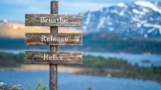 'Breathe. Release. Relax' quote text on wooden signpost outdoors in landscape scenery during blue hour and sunset.