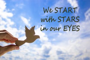 Hand holding wooden bird on lightly cloudy sky background with inspirational quote 'We start with stars in our eyes'