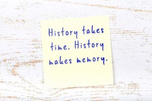 Yellow sticky note with a smart time quote handwritten on it and hanging on wooden wall - 'History takes time. History makes memory.'